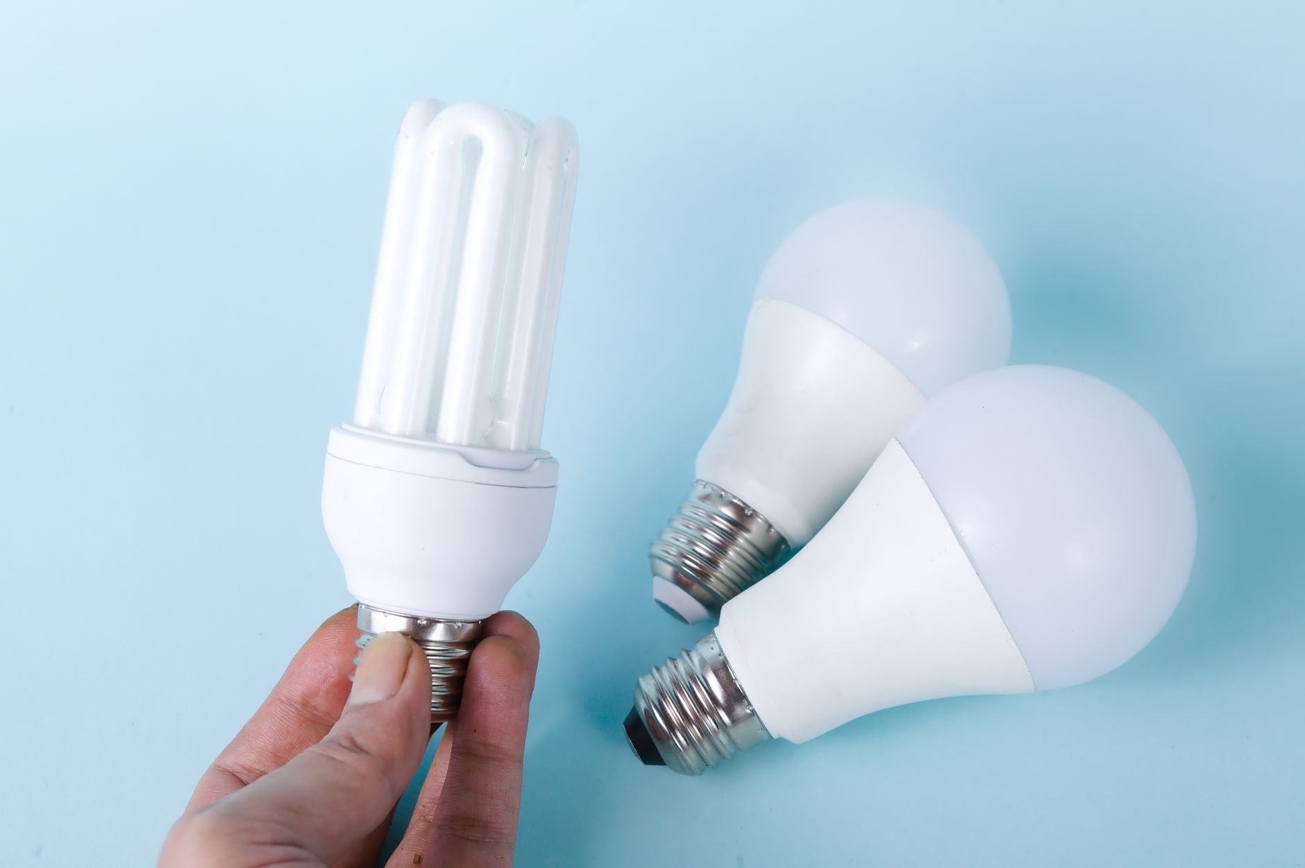 Une main tient une ampoule fluocompacte blanche à côté de deux ampoules LED à bulbe dépoli posées sur un fond bleu clair