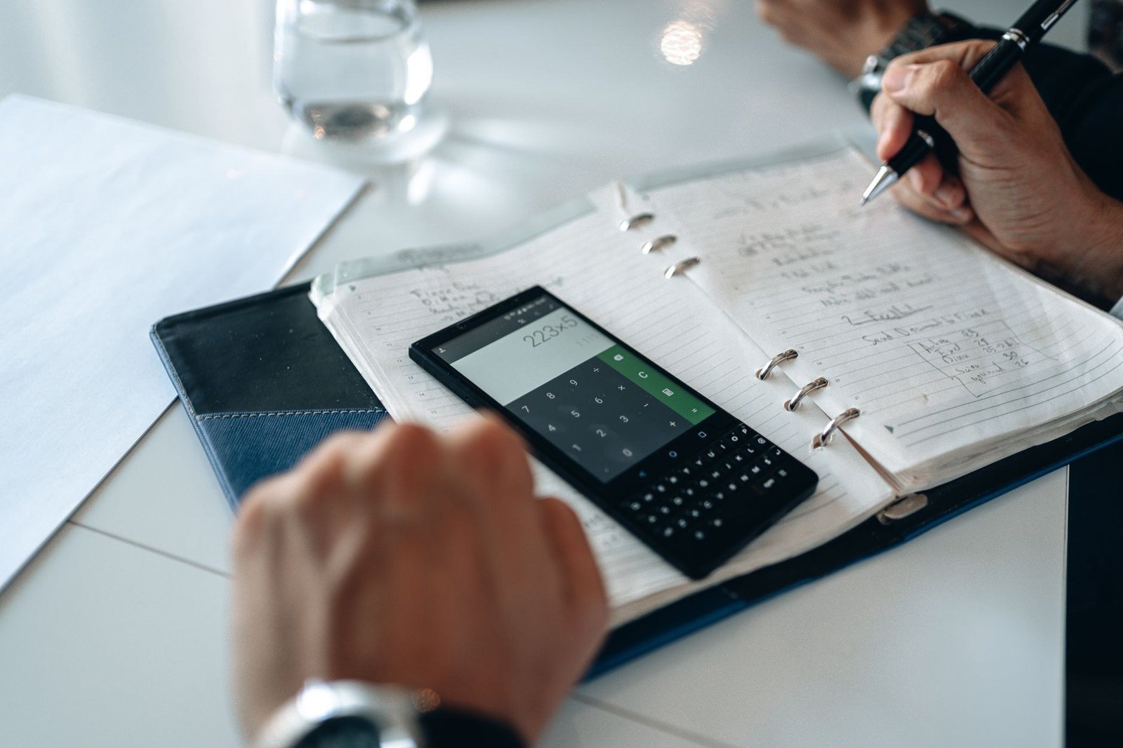 Mains notant des chiffres dans un agenda à anneaux, avec une calculatrice affichée sur un téléphone posé sur la page
