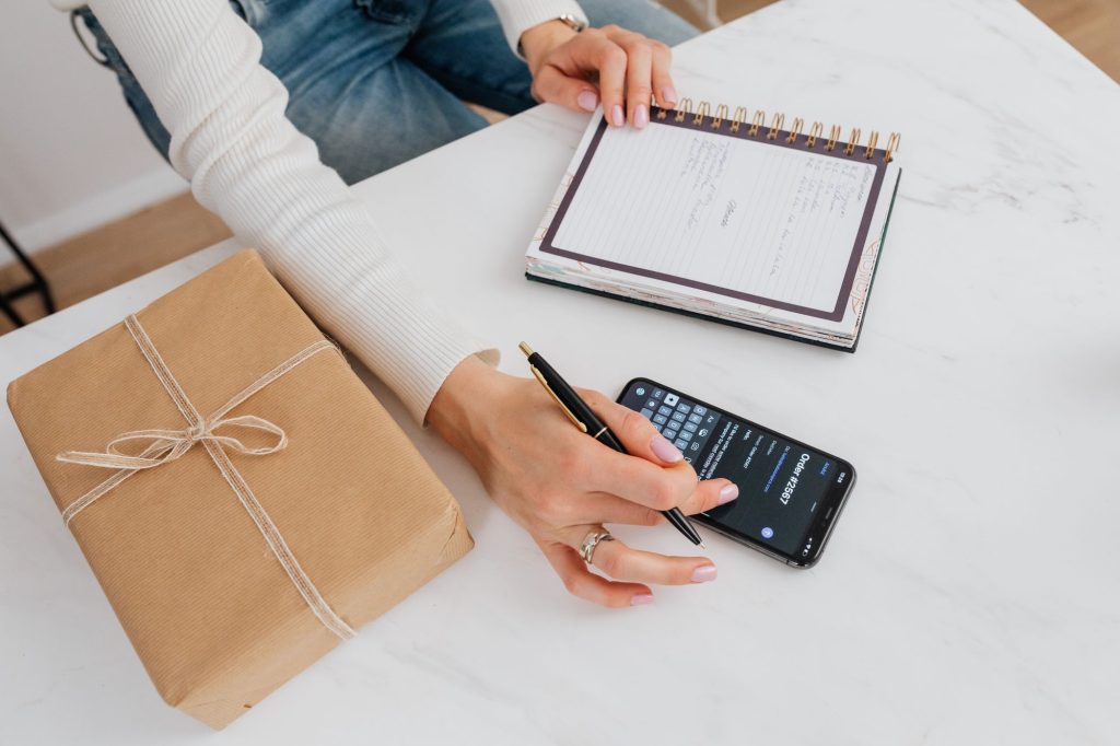 Personne notant son budget dans un carnet à côté d'un smartphone et d'un cadeau emballé