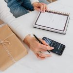 Personne notant son budget dans un carnet à côté d'un smartphone et d'un cadeau emballé