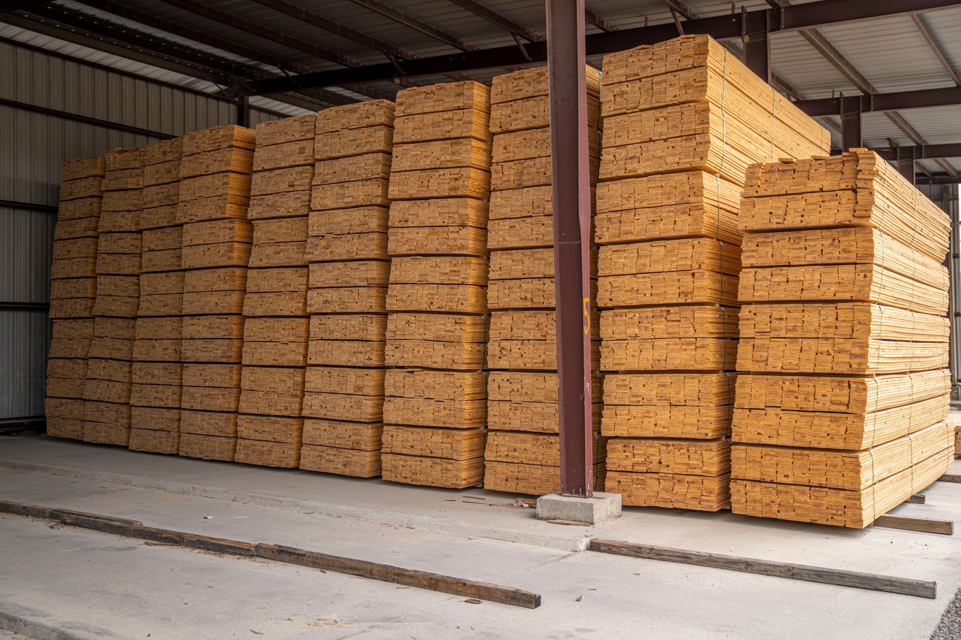 Grandes piles de planches de bois clair empilées dans un entrepôt couvert, séparées par un poteau métallique