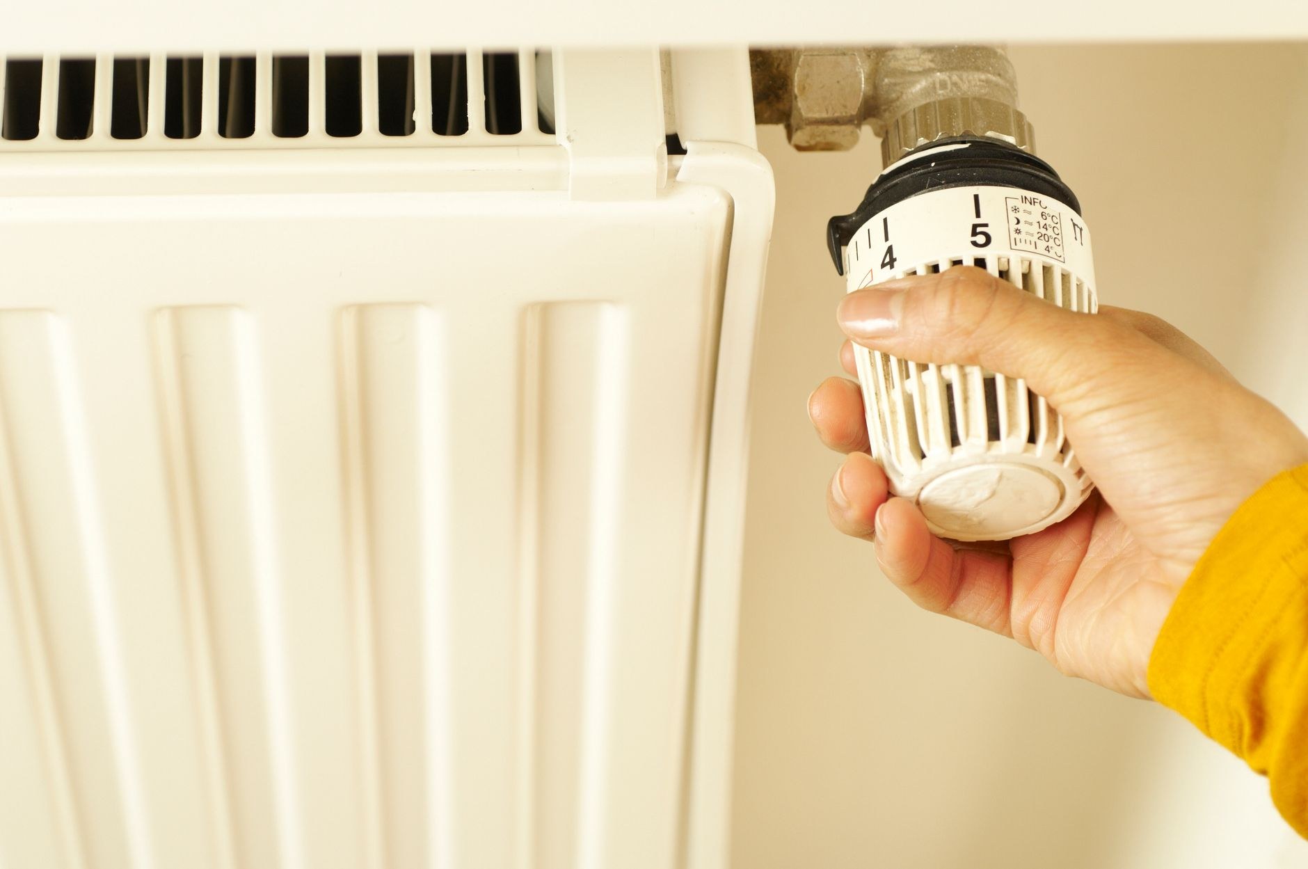 Une main tourne la tête d'un robinet thermostatique, gradué de 1 à 5, sur un radiateur blanc