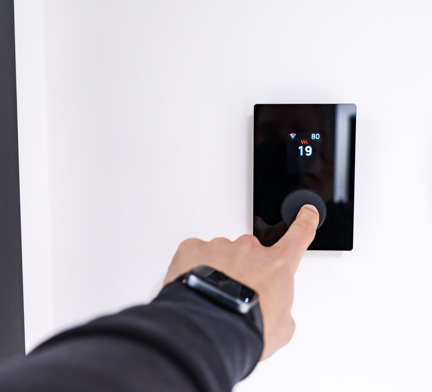 Une main appuie sur un thermostat mural noir qui affiche 19 degrés, sur un mur blanc