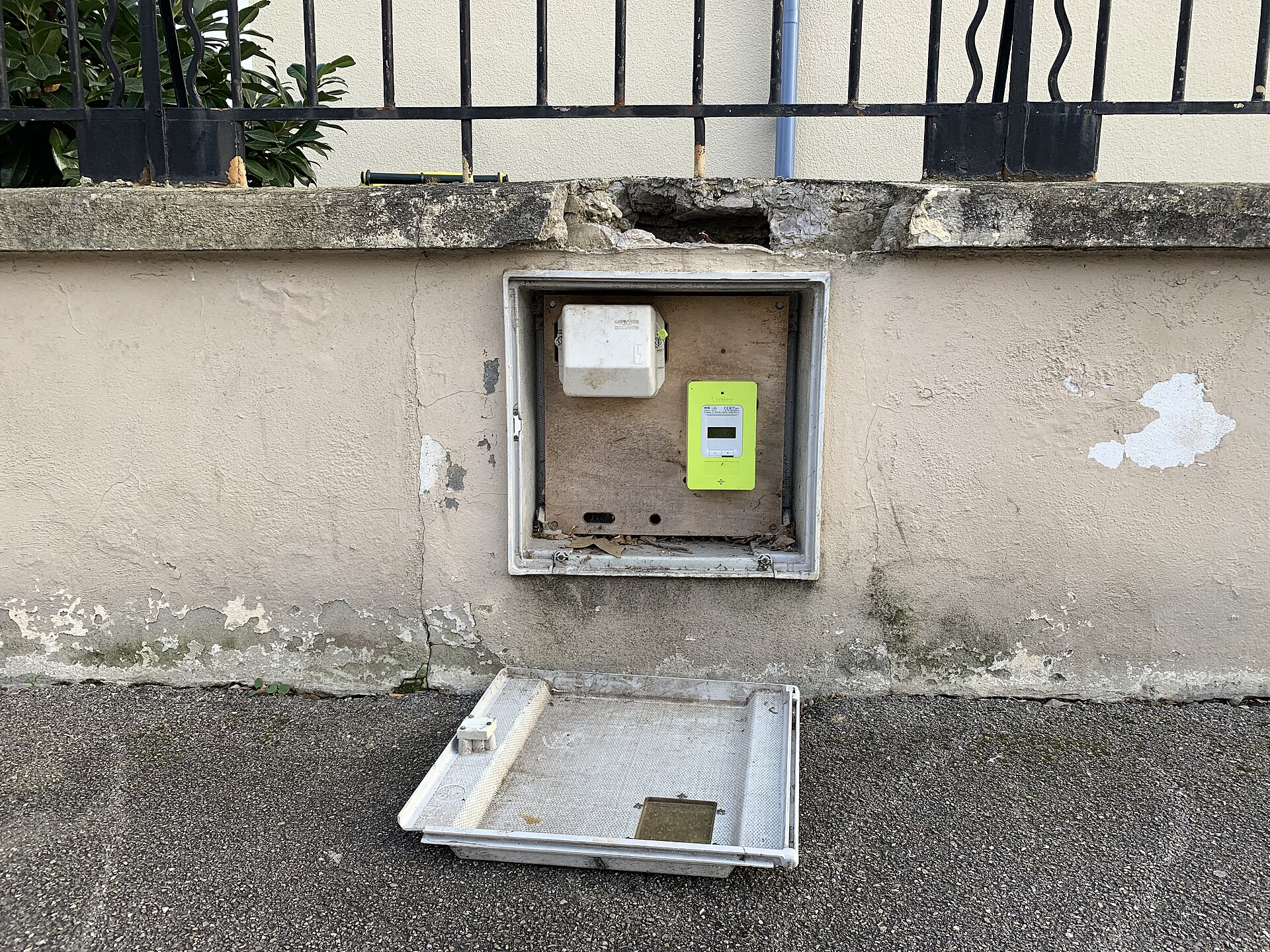 Compteur Linky vert dans un coffret encastré dans un mur de clôture, avec le volet ouvert posé au sol