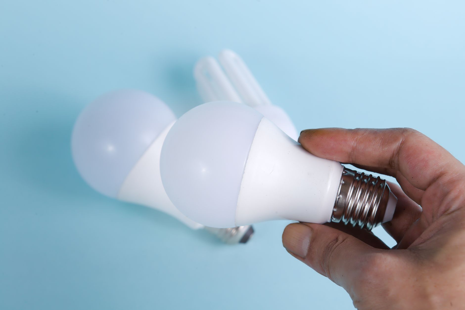 Main tenant une ampoule LED blanche à culot à vis, à côté d’une autre ampoule LED et d’une ampoule fluocompacte