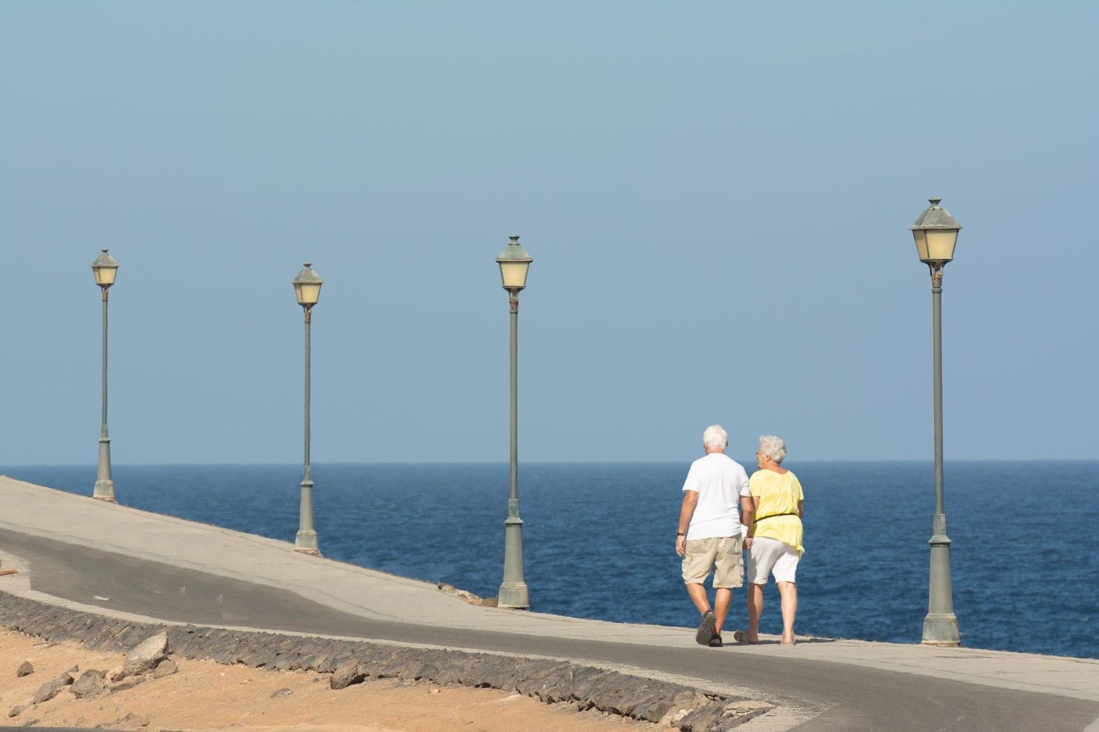 Un couple de personnes âgées vu de dos marche sur une promenade au bord de la mer, entre des lampadaires