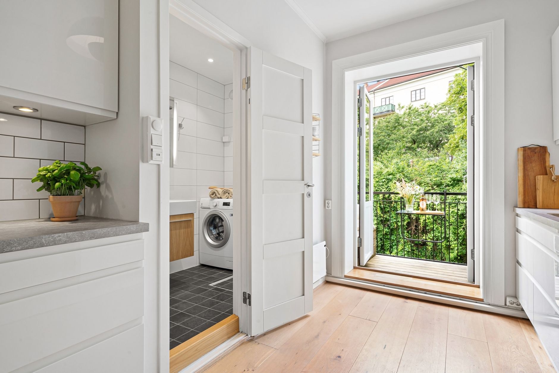 Vue à travers une porte ouverte d’un lave-linge à hublot installé dans une petite salle de bain d’appartement, à côté d’une cuisine claire avec balcon