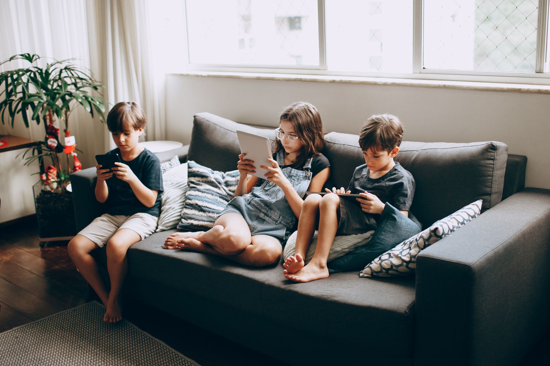 Trois enfants assis sur un canapé gris, chacun absorbé par une tablette ou un smartphone