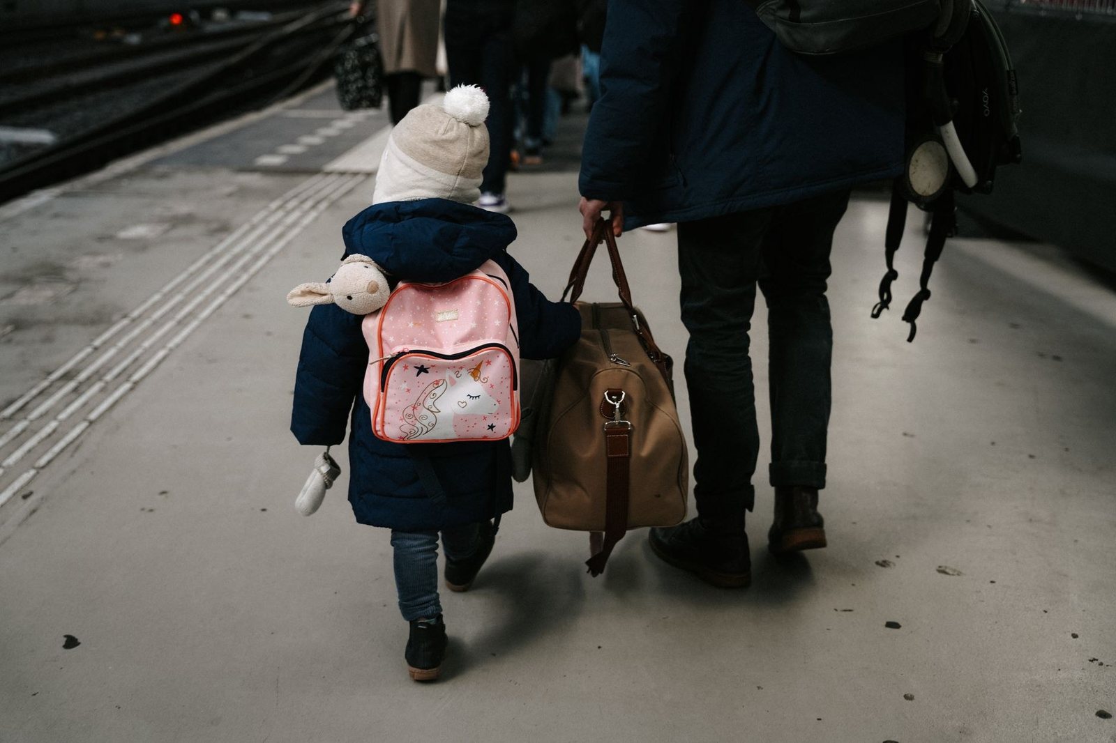 Une famille sur un quai de gare : un jeune enfant avec son sac à dos rose tient la main d’un adulte chargé d’un grand sac de voyage