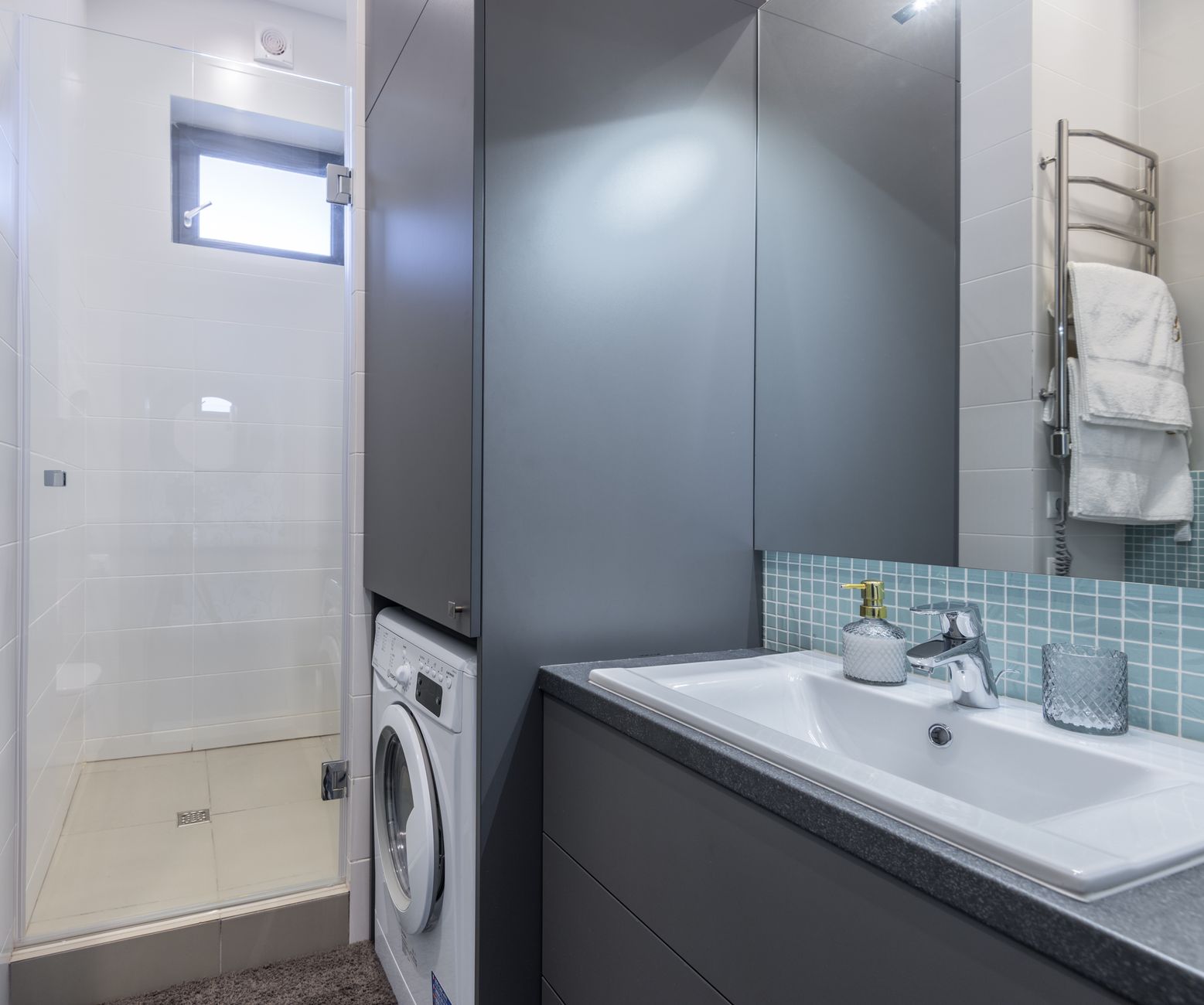 Petite salle de bain avec une douche à paroi vitrée, un lave-linge encastré sous un rangement en colonne et un meuble vasque gris