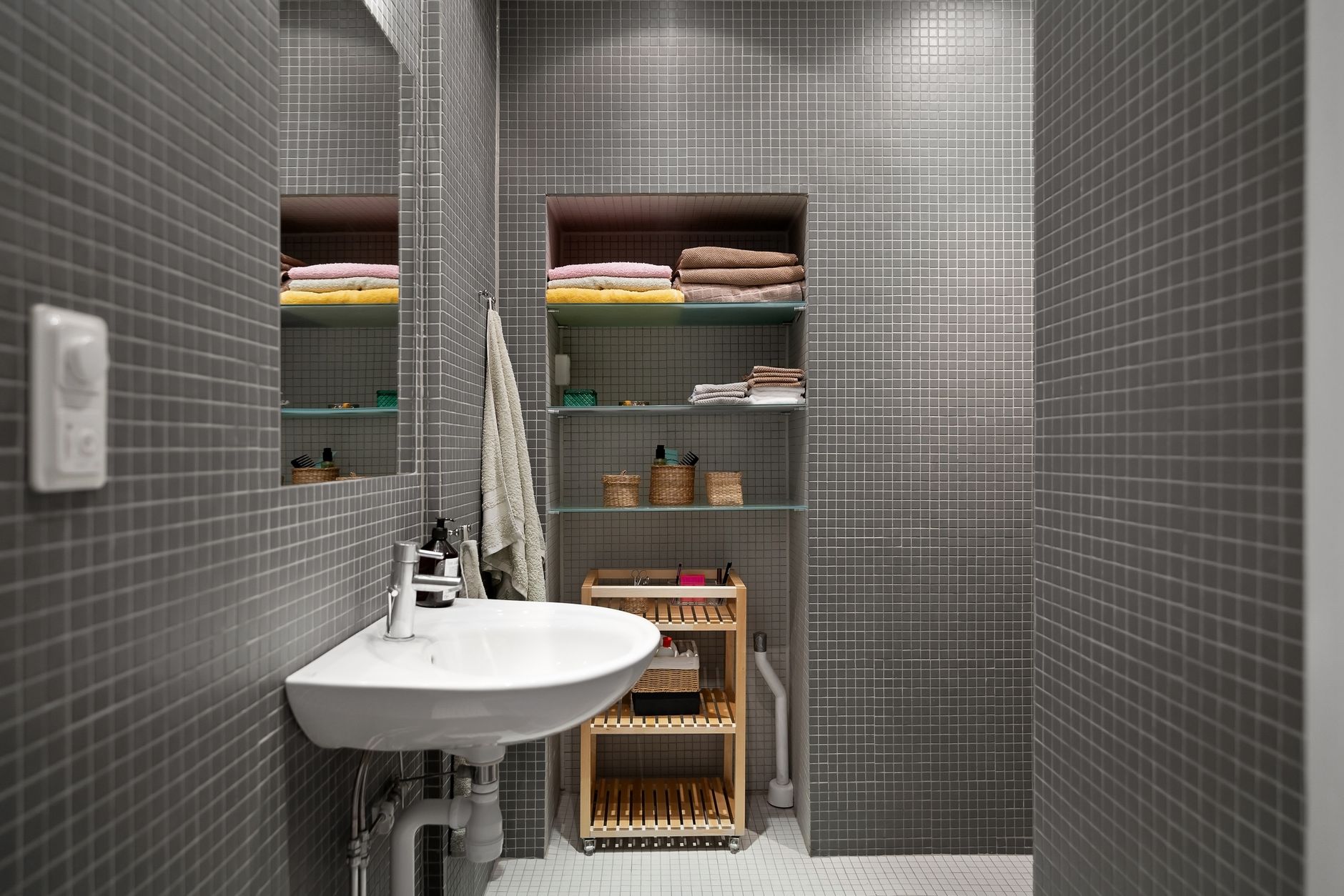 Petite salle de bain carrelée en mosaïque grise avec un lavabo suspendu, des niches à étagères en verre remplies de serviettes et un chariot de rangement en bois