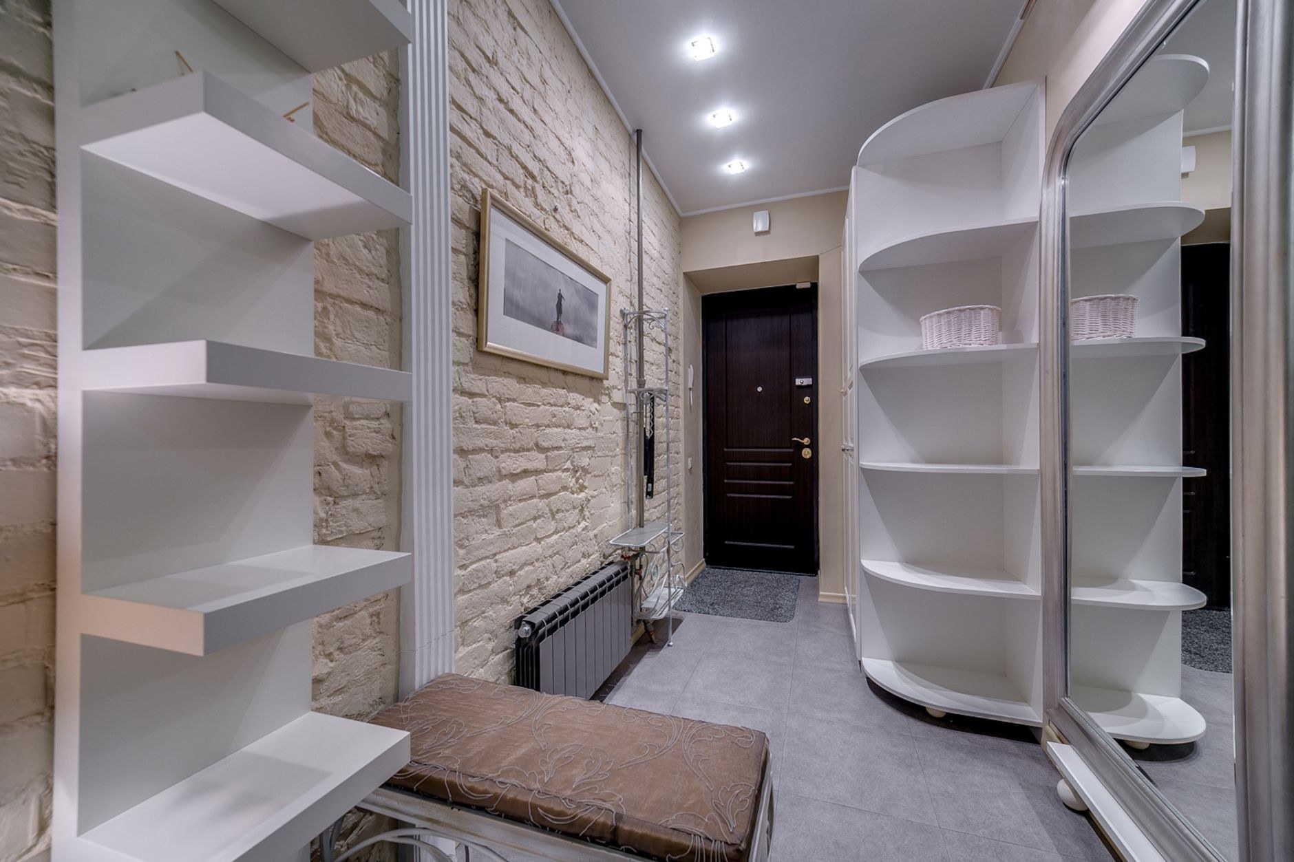 Entrée d'un petit appartement avec des étagères blanches fixées au mur et une étagère d'angle arrondie, près d'un mur en briques claires