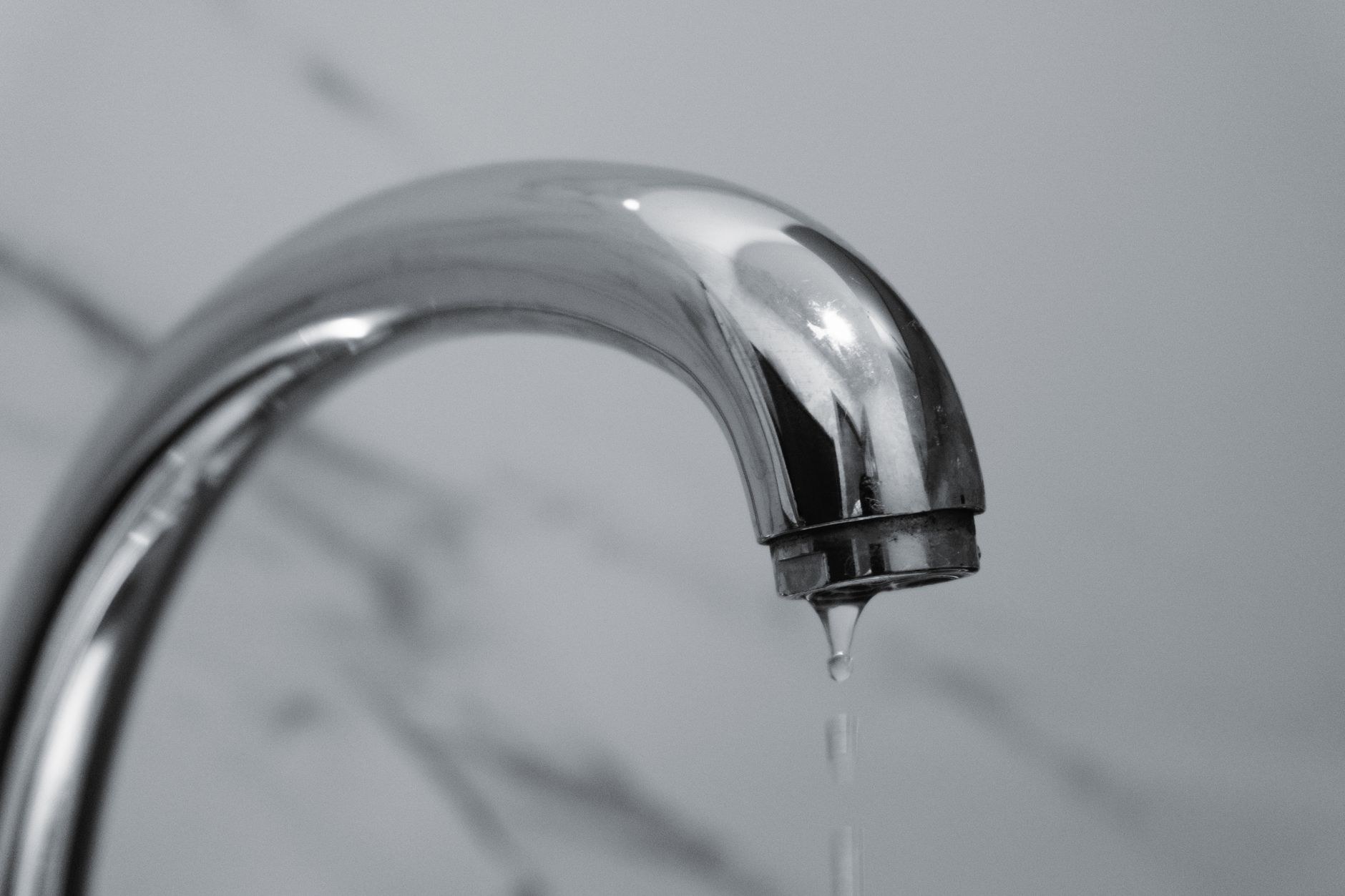 Bec d'un robinet chromé d'où tombe une goutte d'eau, devant un mur gris marbré