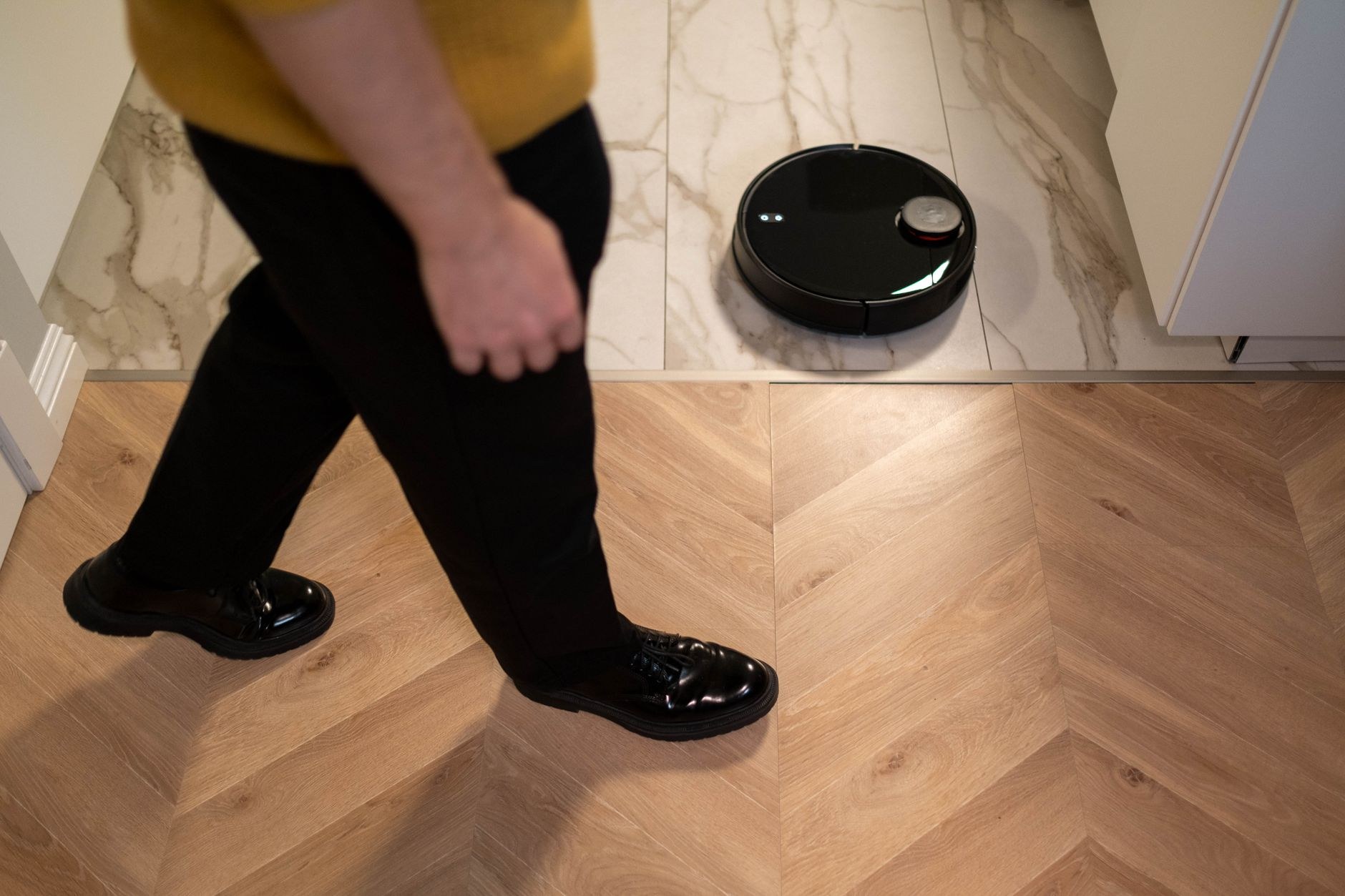 Robot aspirateur noir à capteur circulaire posé sur un carrelage effet marbre, à la jonction avec un parquet en chevrons, pendant qu’une personne passe au premier plan