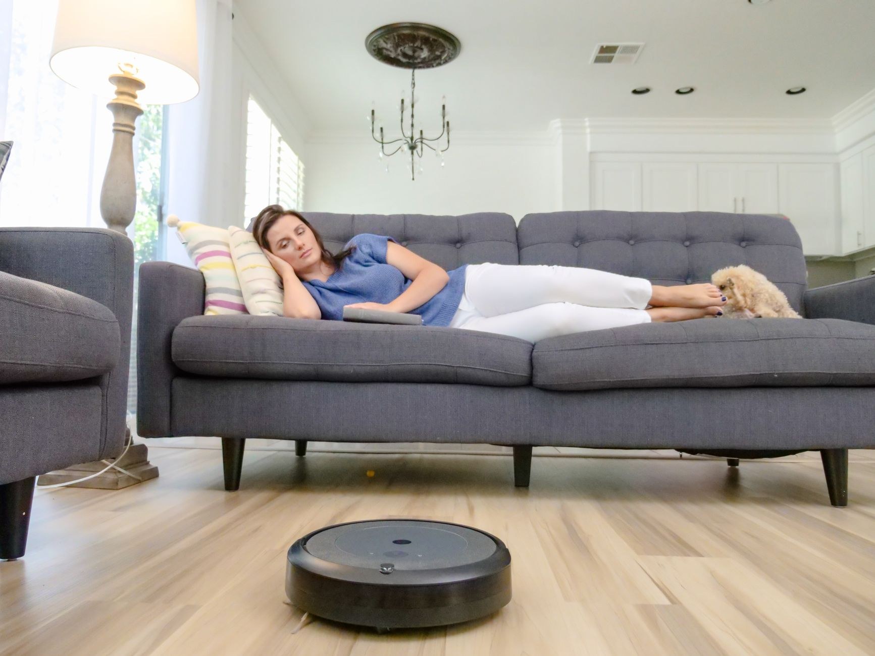 Robot aspirateur rond noir sur un parquet clair devant un canapé gris où une femme est allongée près d’un petit chien