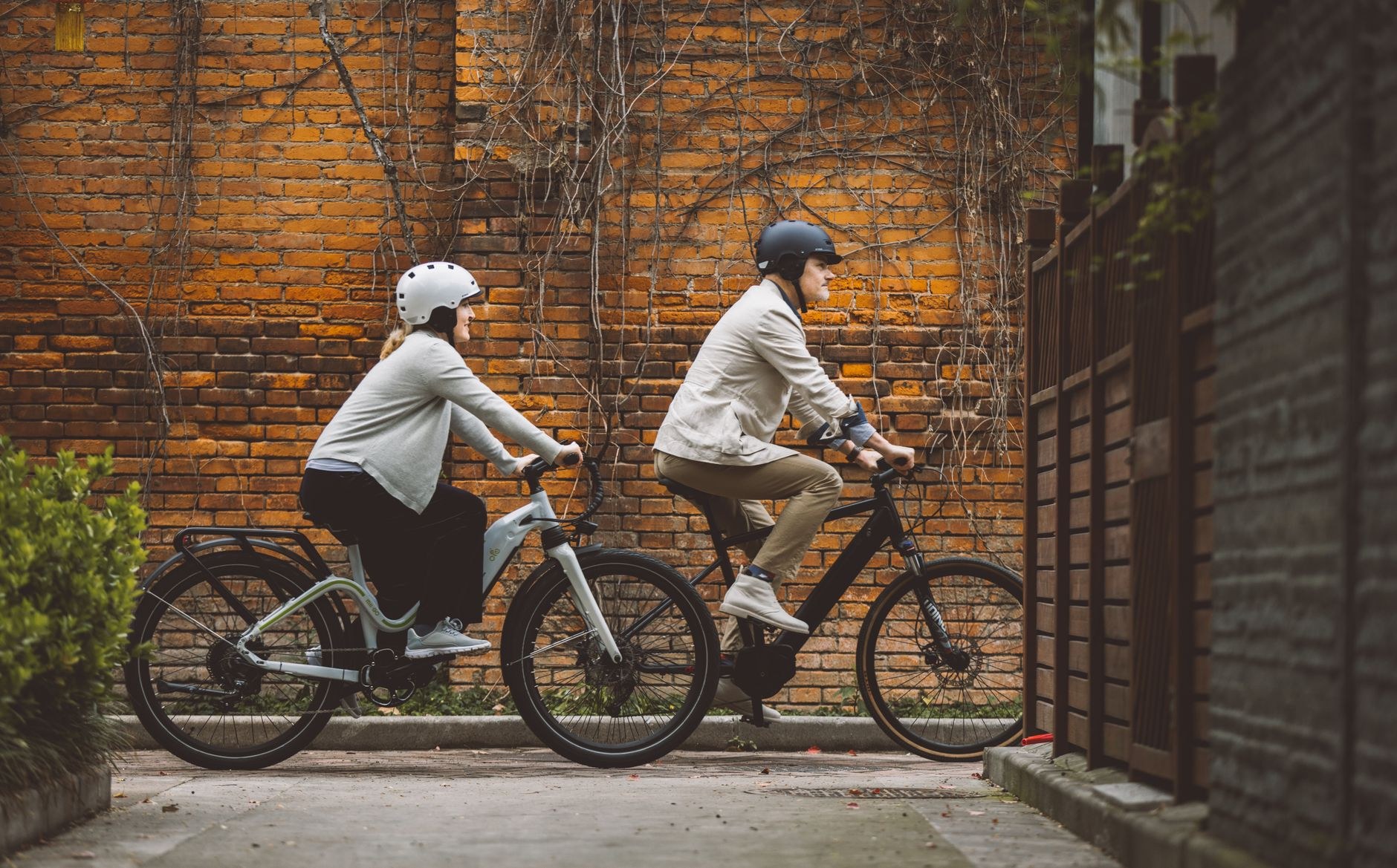 Un homme et une femme casqués roulent côte à côte à vélo électrique devant un mur de briques