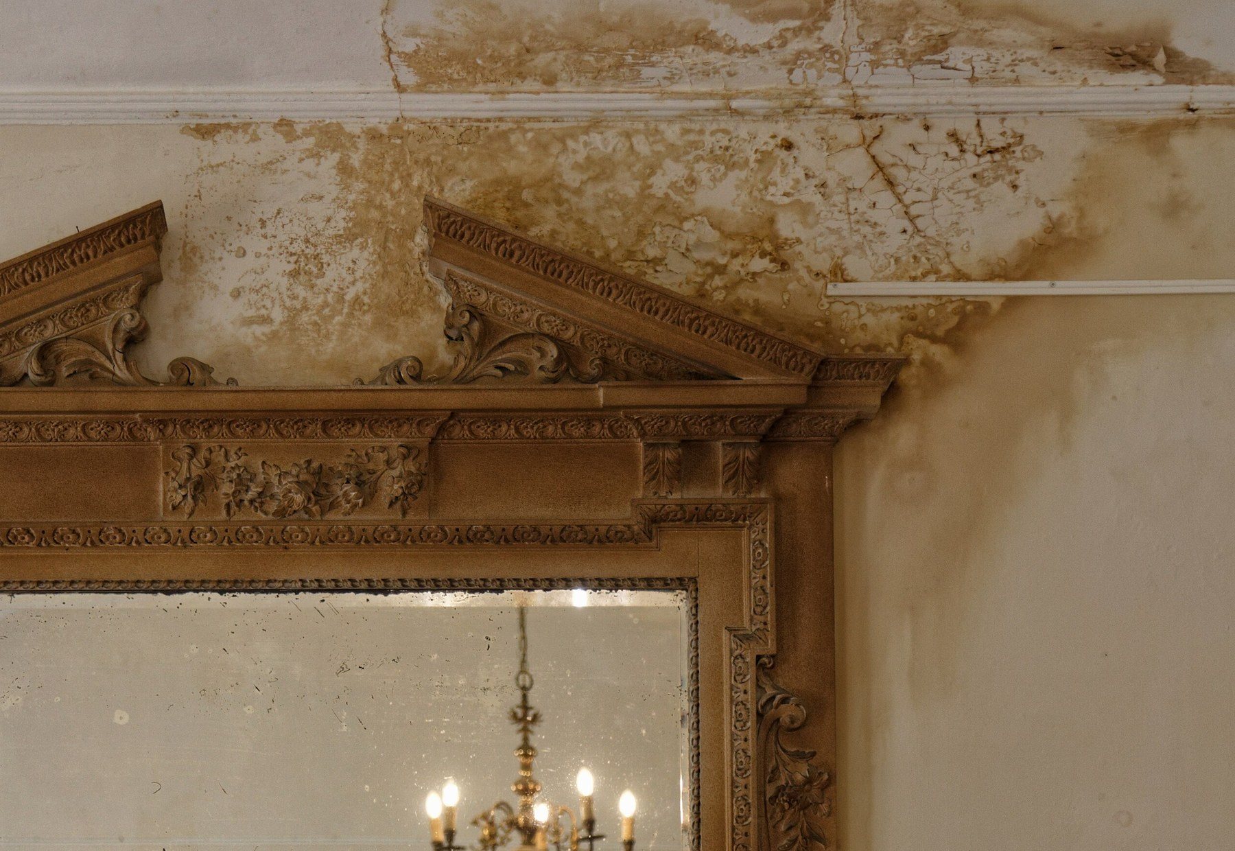 Mur et plafond d'une pièce marqués de larges auréoles brunes et de peinture écaillée après une infiltration d'eau, autour du cadre sculpté d'un miroir