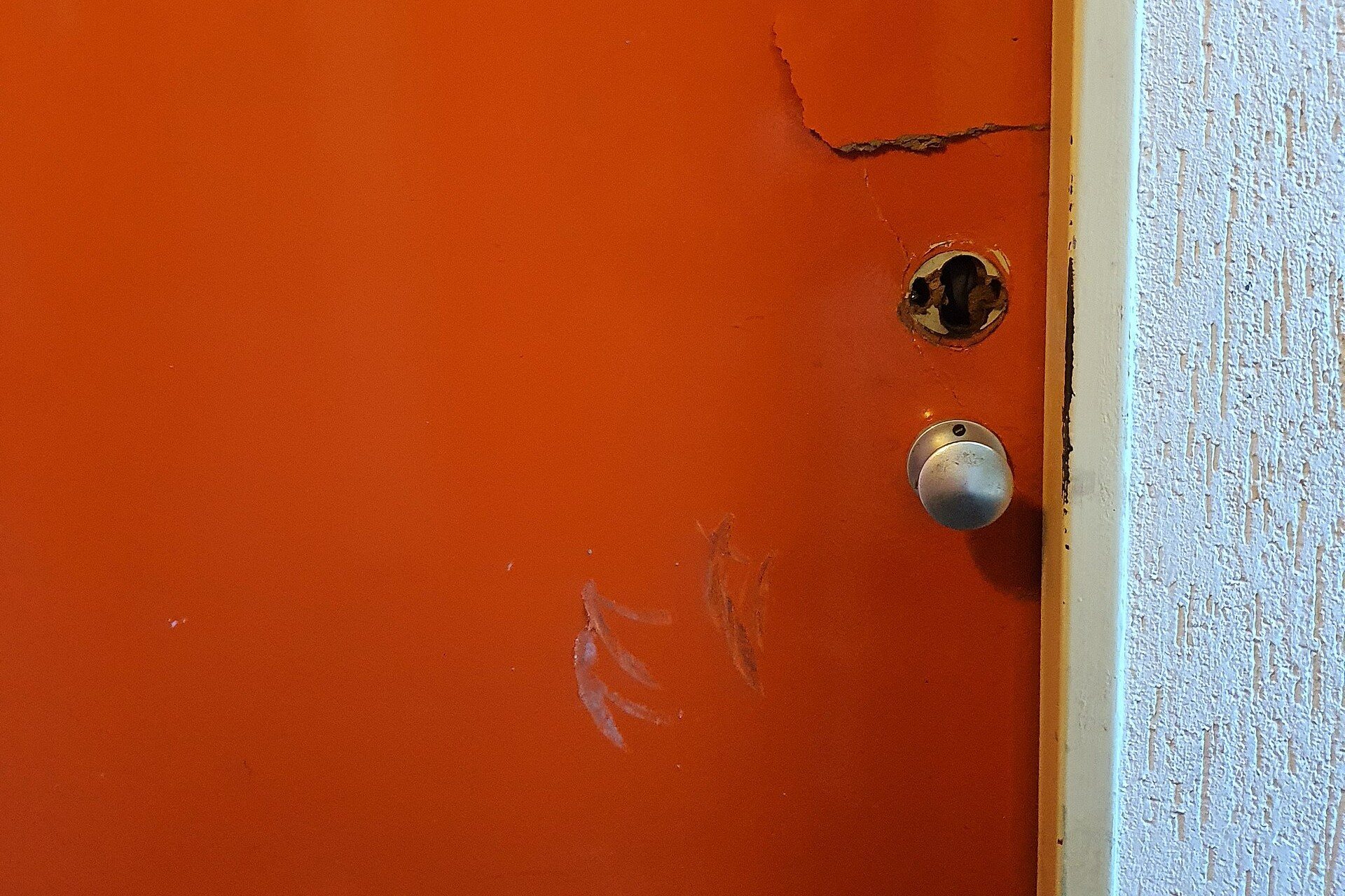 Porte d'entrée orange dont la serrure a été arrachée, laissant un trou dans la serrure, avec la peinture fissurée et des rayures autour de la poignée