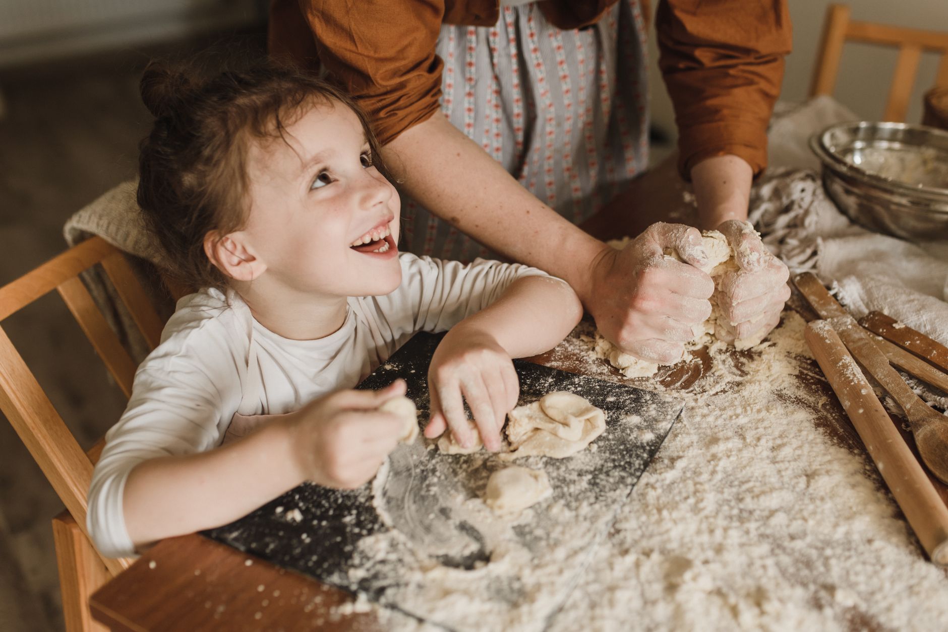 Une petite fille rit en levant les yeux vers l'adulte qui pétrit une pâte farinée sur une table en bois