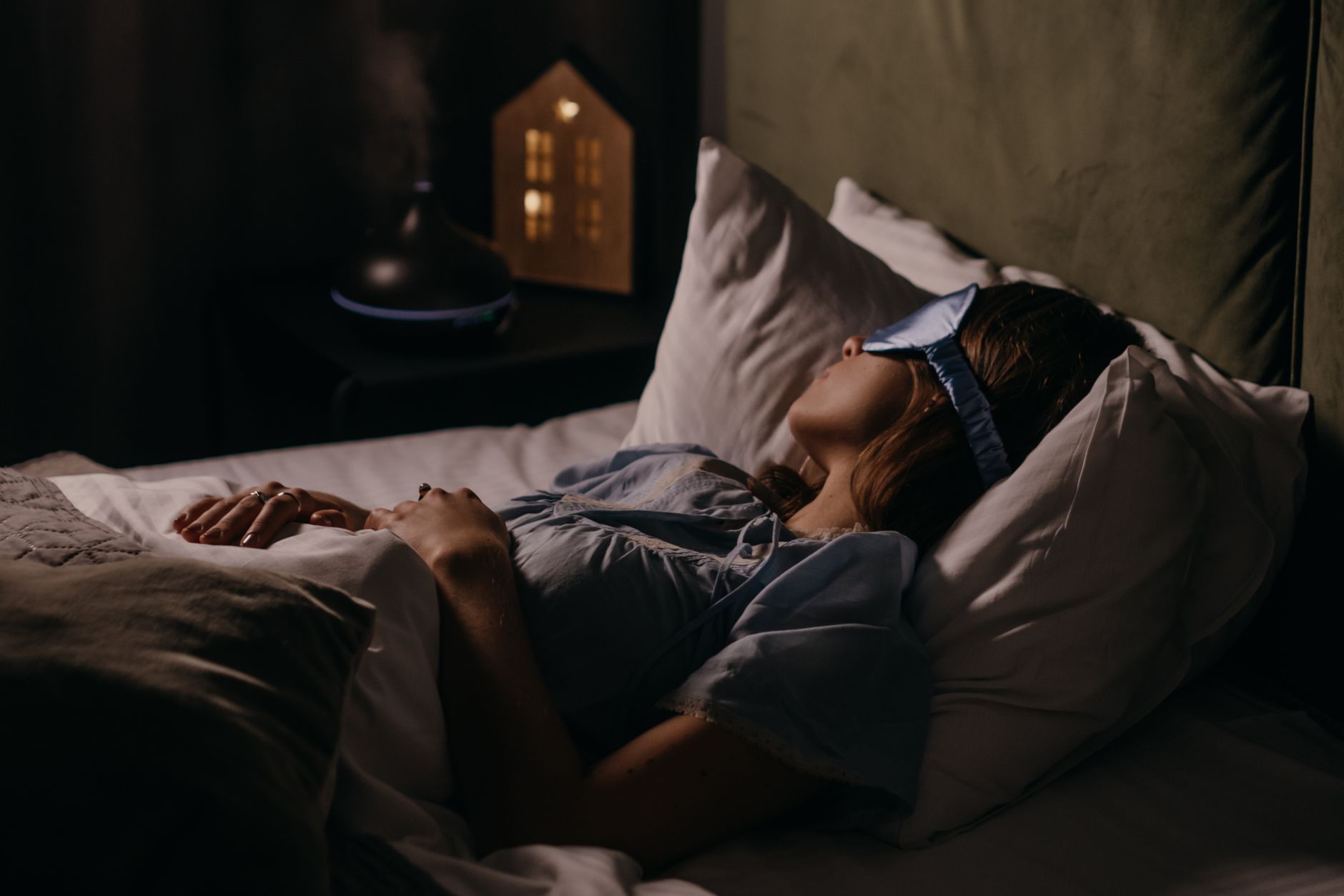 Une femme dort sur le dos dans une chambre plongée dans la pénombre, un masque de nuit bleu sur les yeux, une veilleuse en forme de maison sur la table de nuit
