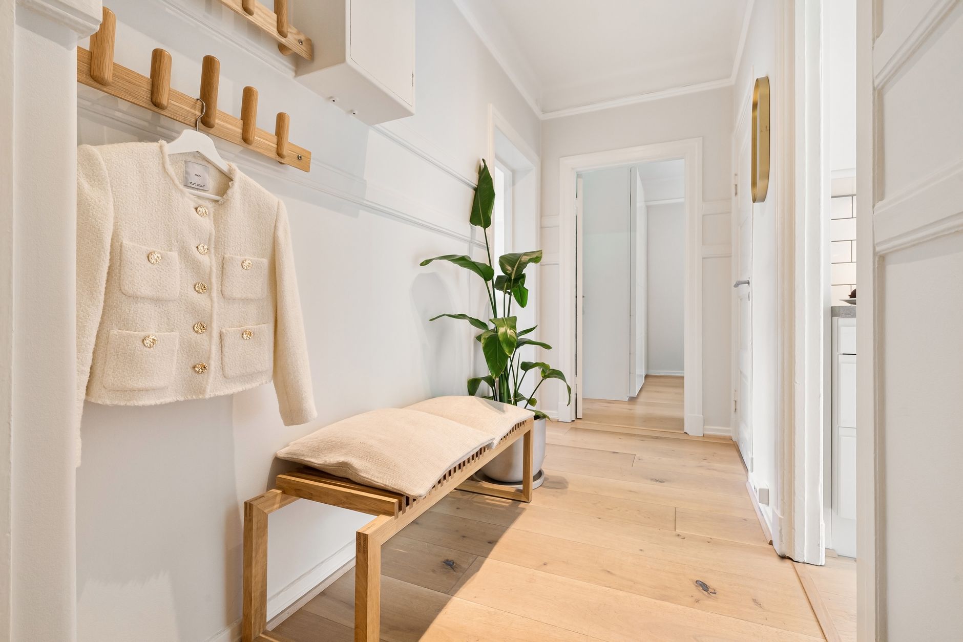 Entrée claire avec un banc en bois et un coussin, une rangée de patères en bois où est suspendue une veste crème, et une plante