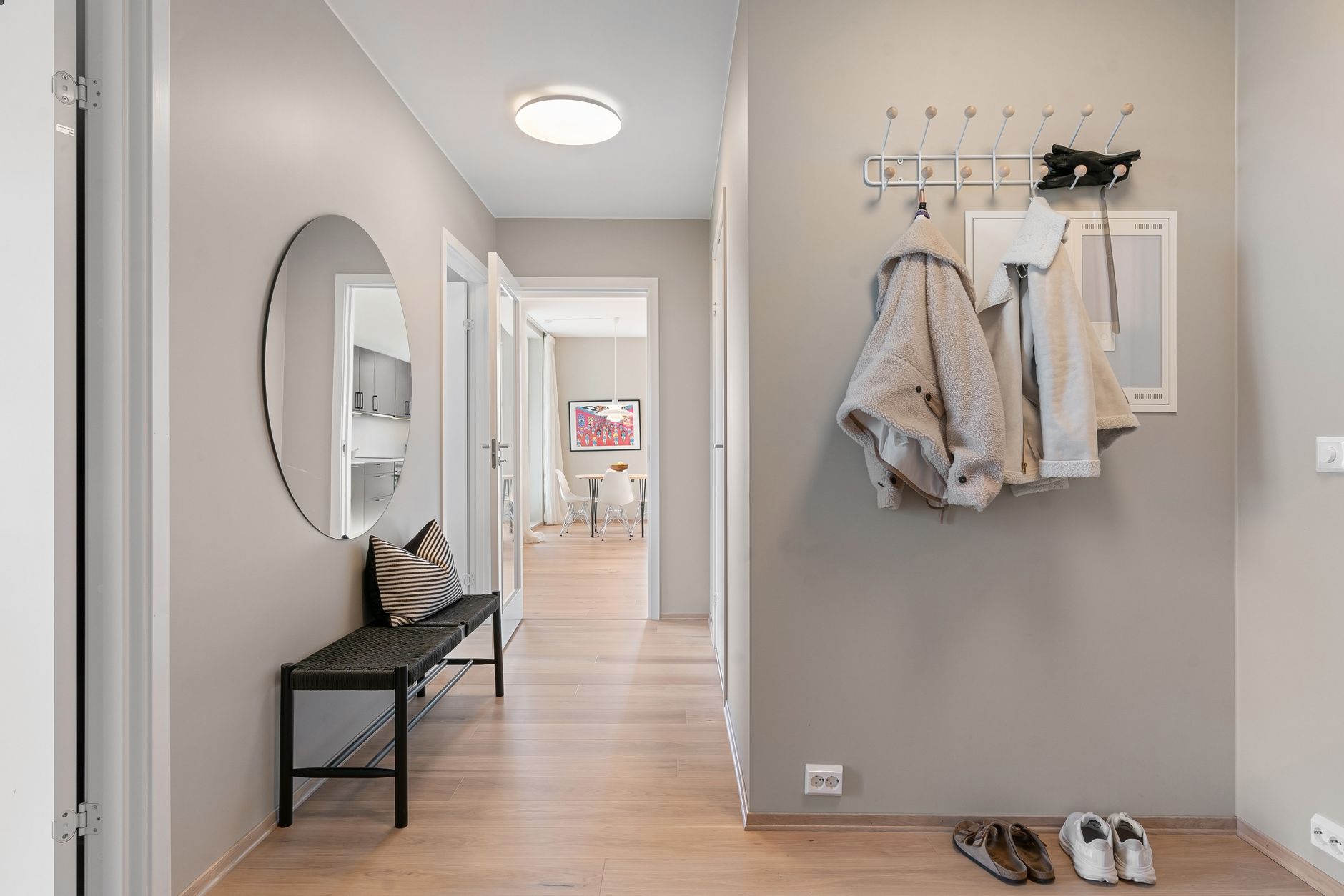 Couloir d’appartement avec un miroir rond, un banc bas noir, une patère murale chargée de manteaux et des chaussures au sol