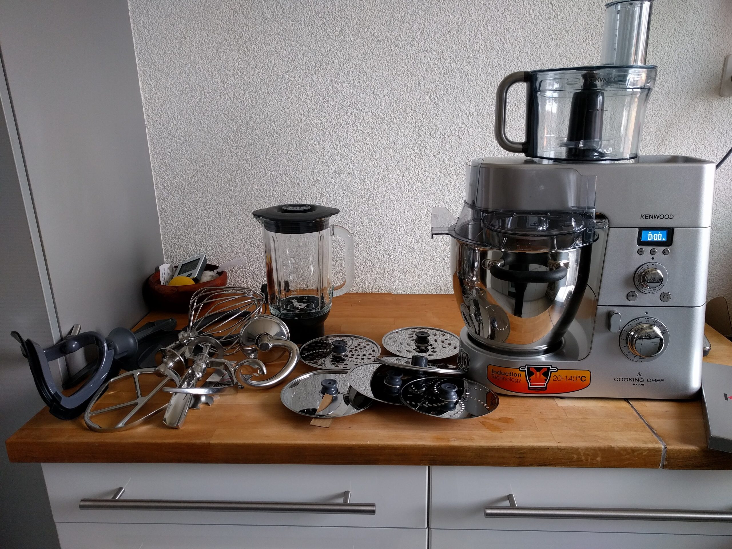 Robot Kenwood Cooking Chef sur un plan de travail en bois avec ses fouets, disques râpeurs et son blender