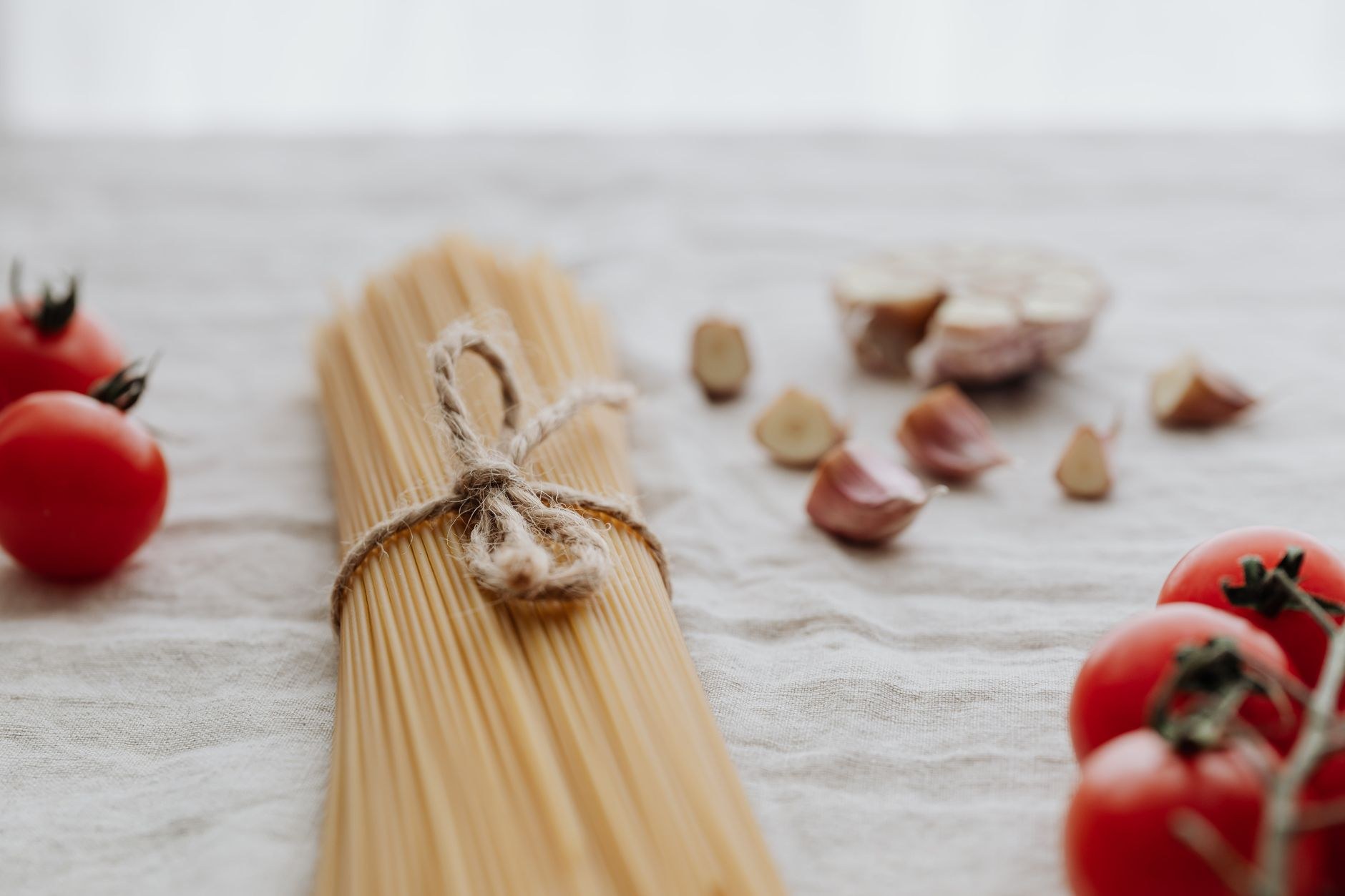 Une botte de spaghettis nouée d'une ficelle, entourée de tomates sur la vigne et de gousses d'ail sur une nappe en lin