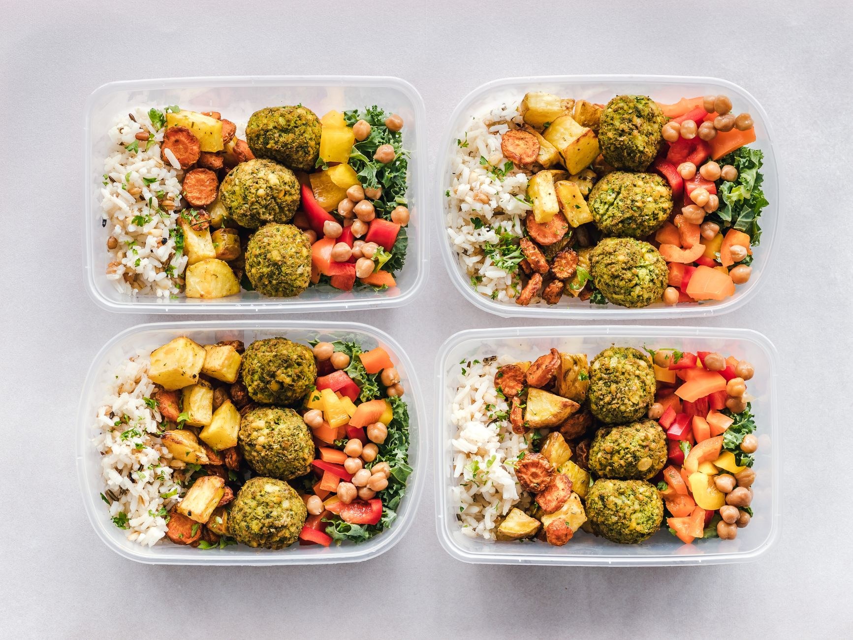 Quatre barquettes transparentes de repas préparés : riz, boulettes vertes, légumes rôtis et pois chiches