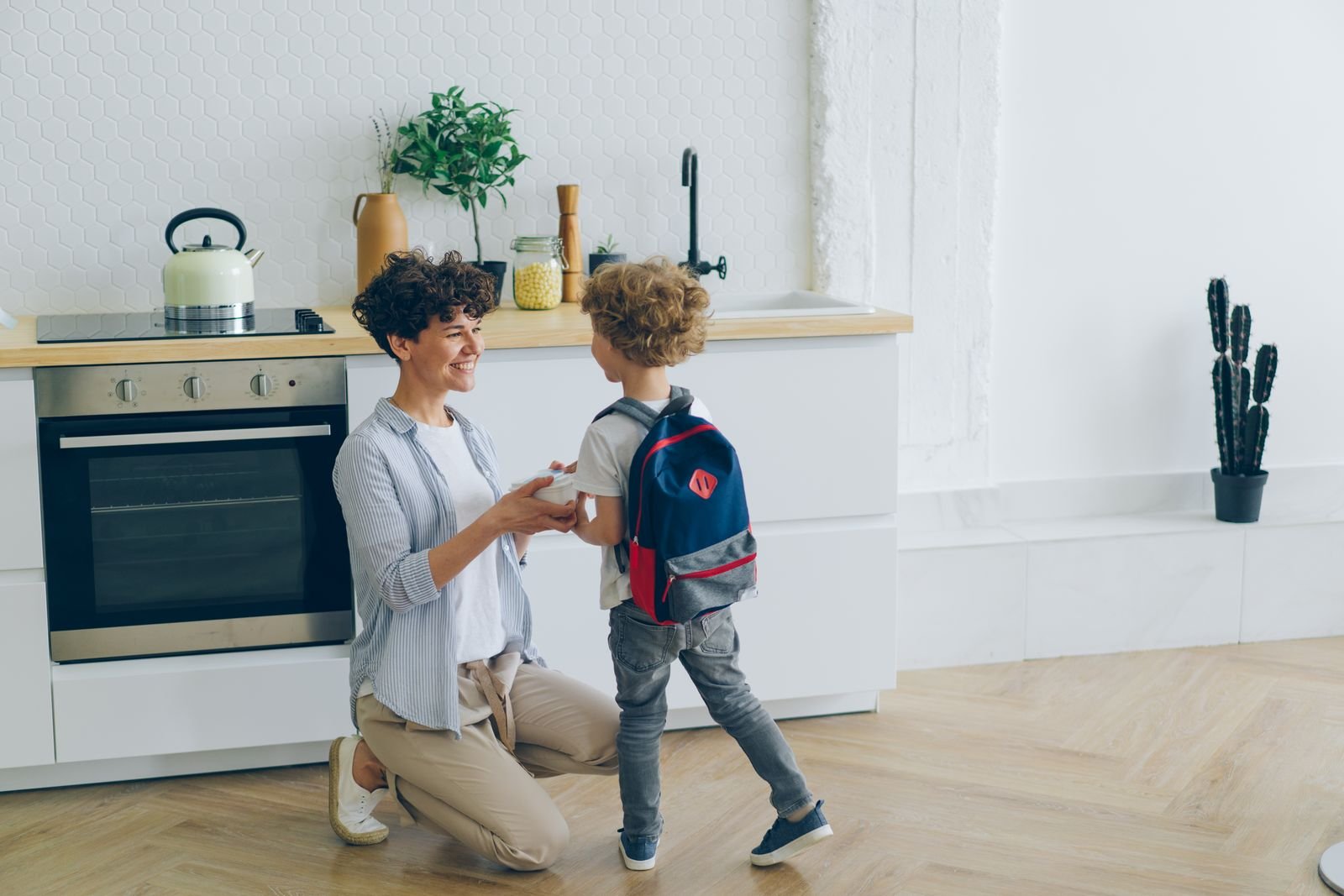 Une mère accroupie ajuste le sac à dos de son fils dans une cuisine avant le départ pour l'école