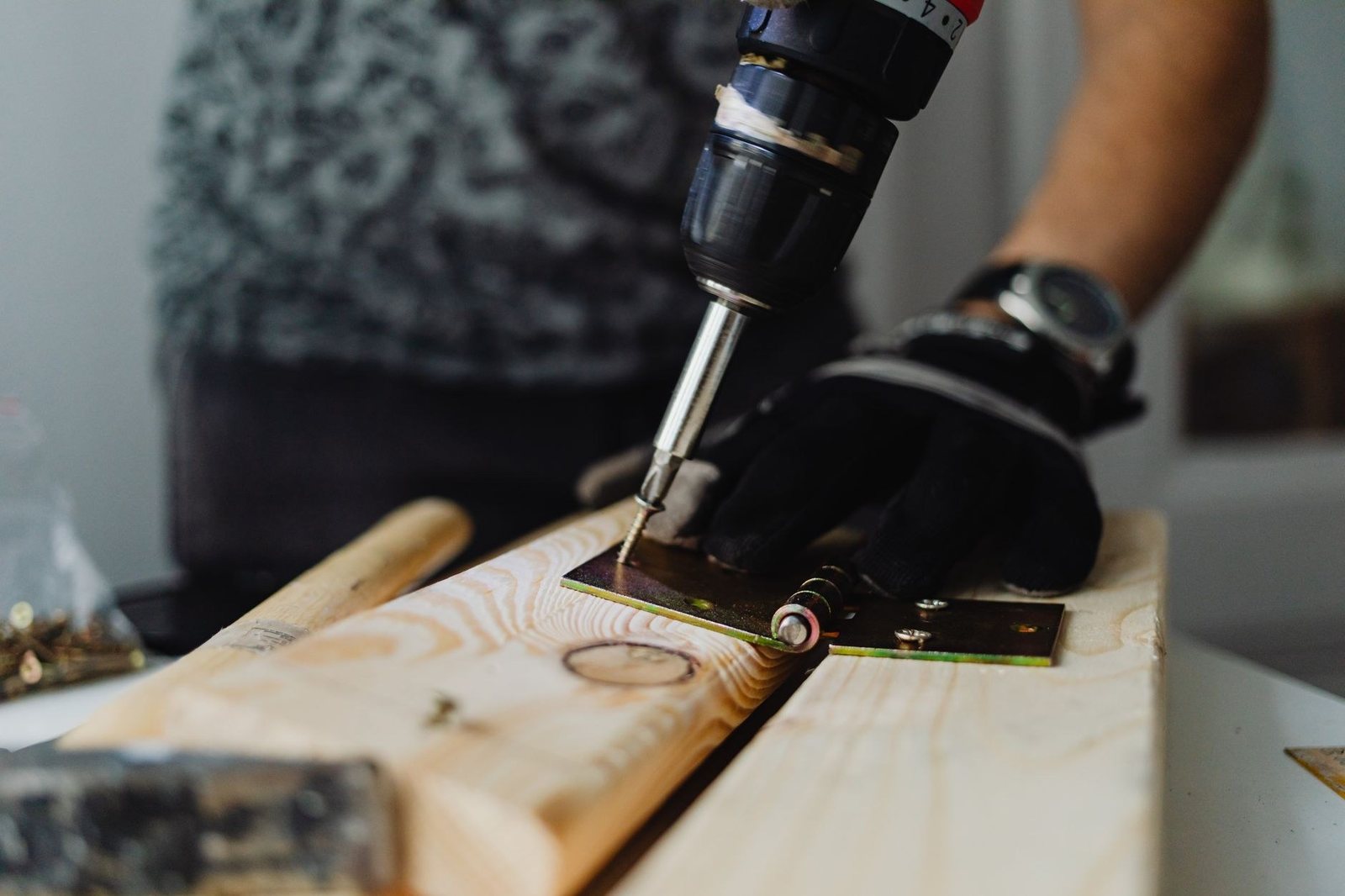 Une main gantée tient une charnière sur une planche de bois pendant qu’une perceuse-visseuse enfonce une vis