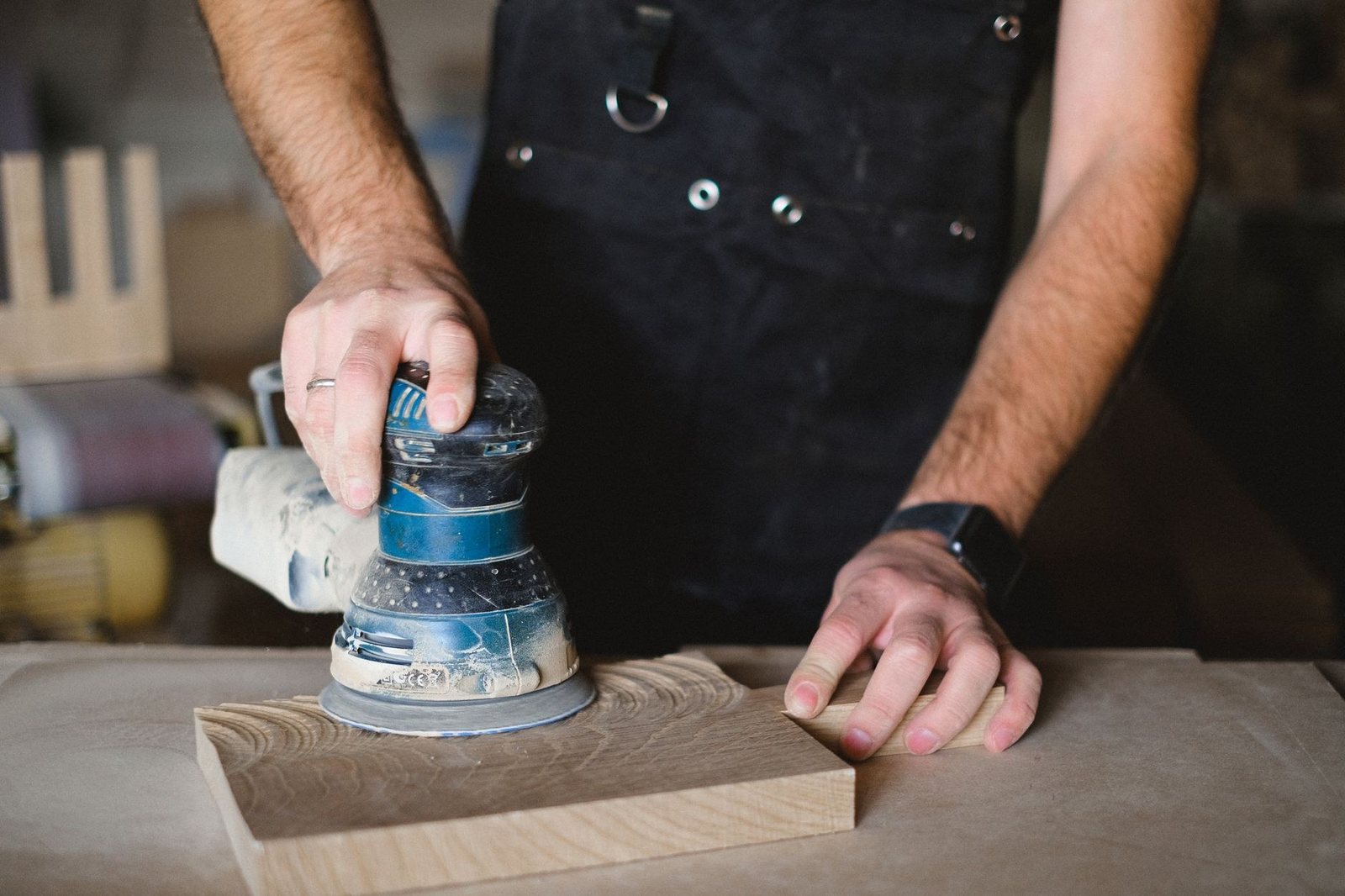 Un artisan ponce une planche de bois avec une ponceuse orbitale bleue, une main posée sur la planche