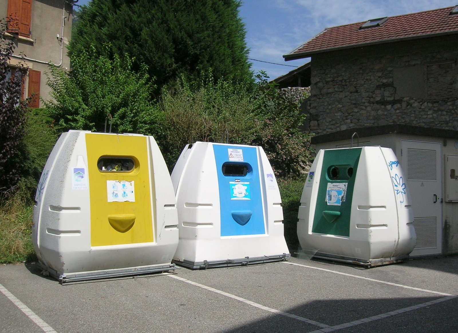 Trois conteneurs de collecte sélective, jaune, bleu et vert, installés sur un parking de village